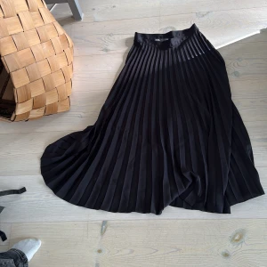Svart plisserad långkjol från Zara - Säljer en svart plisserad långkjol från Zara. Kjolen har snygga, breda veck och en hög midja som ger en stilren och trendig look. Perfekt att matcha med en croppad topp eller stickad tröja för en cool outfit.