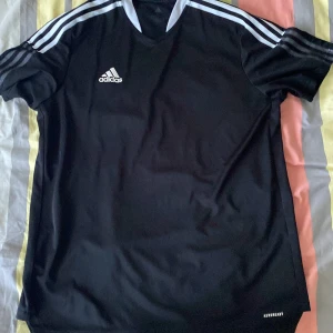 Svart Adidas fotbollströja XL - Snygg svart fotbollströja från Adidas i storlek XL, tillverkad i Primegreen-material. Tröjan har klassiska vita ränder på axlarna, vit V-ringad krage och Adidas-logga på bröstet. Perfekt för träning eller match, med korta ärmar och lätt, ventilerande tyg.