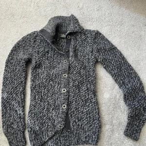 Grå stickad kofta Peak Performance - Säljer en chunky gråmelerad stickad kofta från Peak Performance i storlek XS. Koftan har grovstickad struktur, hög krage och snygga metallknappar med logga. Perfekt för kyliga dagar och har långärmad passform. Materialet är mjukt och värmande ull.