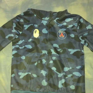 BAPE x PSG camo bomberjacka - Säljer en fet bomberjacka från BAPE x PSG med blått och svart camo-mönster, broderade bokstäver på ärmen och ikoniska haj- och tigertryck. Jackan har dragkedja, huva och stora BAPE-loggan på ryggen. Perfekt för dig som vill sticka ut med streetwear.