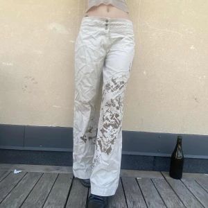 Beiga wideleg byxor med brunt mönster - Säljer ett par beiga wide byxor med brunt abstrakt mönster på ena benet och höften. Byxorna har hög midja med tre knappar framtill och raka ben. Skitcoolt, unikt mönster. Från märket repeat. Midja tvärs över: 41cm, längd 102cm, bredd längst ner på benet 30cm