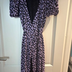 Långklänning med lila prickar - Snygg långklänning med svart botten och lila prickigt mönster från Cubus. Klänningen har omlottmodell med v-ringad hals, korta puffärmar och ett knytband i midjan. 