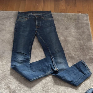 Blå jeans från Nudie Jeans grim tim  - Säljer ett par klassiska blå jeans från Nudie Jeans med snygga orangea sömmar och deras ikoniska bakficka med broderad våg. Jeansen har rak passform och normal midja, tillverkade i slitstarkt bomullsmaterial. Perfekt för dig som gillar tidlös stil och streetwear.