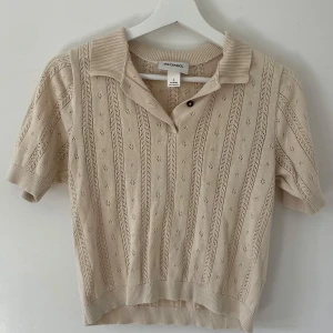 Stickad blus  - Säljer en beige stickad blus från Monki i storlek S. Tröjan har korta ärmar, krage och knappar framtill. Den är dekorerad med ett snyggt hålmönster och ribbade muddar vid ärmslut och nederkant. Perfekt för vår och sommar!