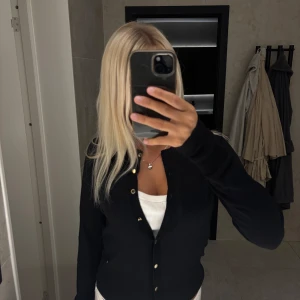 Svart kofta från Holly & Whyte - Svart kofta  från Holly & Whyte by Lindex. Den har guldfärgade knappar framtill och är långärmad med ribbade muddar. Materialet är mjuk bomull och koftan har en clean och klassisk look.