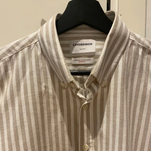Randig och grön skjorta från Lindbergh - Två skjortor från Lindbergh i slim fit. Ena är beige och vit med vertikala ränder, klassisk krage och knappar framtill. Den andra är enfärgad ljusgrön med samma stilrena design och knappar.                                              450kr/st, 800kr för båda