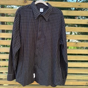 Svart rutig skjorta från Bläck XXL - Svart skjorta från Bläck i storlek XXL med vitt rutmönster. Tillverkad i 100% bomull, har klassisk krage, långa ärmar och knäppning framtill. Perfekt för dig som gillar stilrena och grafiska mönster.