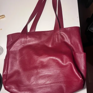 Stor vinröd vintage skinnaxelväska - Snygg röd axelväska i skinnimitation med två axelband. Väskan har ett rymligt huvudfack, innerfack med dragkedja och en öppen ficka. Perfekt för att få plats med allt du behöver och samtidigt hålla stilen. Perfekt skolväska, får plats med allting. Jättesnygg vintage väska.