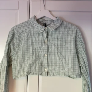 Grön rutig croppad skjorta - Croppad skjorta från H&M Divided i ljusgrönt och vitt rutigt mönster. Skjortan har klassisk krage, långa ärmar med knapp vid manschetten och knäppning framtill.