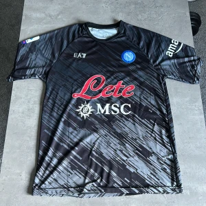 Napoli Kvaratskhelia matchtröja - Napoli fotbollströja med Kvaratskhelia och nummer 77 på ryggen. Storleken är XL men sitter även bra för de som har strolek L. Svart bas med grått grafiskt mönster, blått klubbmärke och vita samt röda tryck från Lete, MSC och Amazon. Kortärmad, rund hals och tillverkad i lätt polyester för max komfort.
