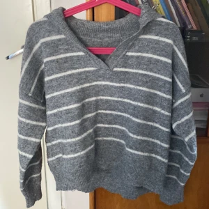 Grå randig stickad tröja med v-ringning - Mysig grå stickad tröja med vita ränder och v-ringad hals. Tröjan har långa ärmar och ribbade muddar vid ärmslut och nederkant. Perfekt för kyliga dagar och passar till jeans eller kjol. Snygg och enkel design som är lätt att matcha.