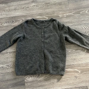 Mörkgrå stickad kofta från 157 - Mysig mörkgrå stickad kofta från 157 i storlek XS. Koftan har rund hals, långa ärmar och knappar hela vägen framtill. Perfekt att slänga på sig när det är lite kyligt ute. Enkel och stilren design som funkar till många olika outfits.