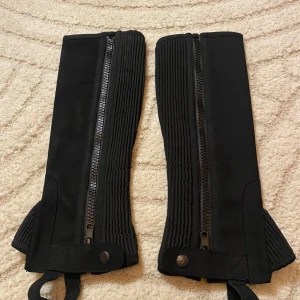 Svarta ridstövelskydd - Säljer ett par helt oanvända ridstövelskydd ( half chaps) med super grip. Svarta snygga och funktionella med dragkedja. 