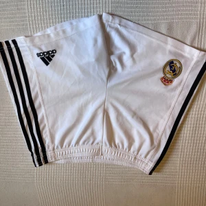 Vita Real Madrid shorts från Adidas - Snygga vita Real Madrid shorts från Adidas i storlek M med svarta ränder längs sidorna och broderad klubbmärke. Elastisk midja för skön passform. Perfekta för fotboll eller chill. Materialet är polyester som andas bra.