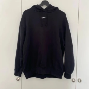 Svart hoodie från Nike med huva - Svart hoodie från Nike med klassisk vit swoosh på bröstet. Tröjan har huva med snörning och långa ärmar. Mjuk och skön bomullsmix som passar perfekt när du vill ha en chill och enkel stil. Oversized passform för extra comfort.