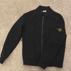 Svart bomberjacka från Stone Island - Snygg svart bomberjacka från Stone Island Junior med klassisk logotyp på ärmen. Jackan har rak passform, lång ärm och dragkedja framtill. Materialet känns mjukt och har en clean look, perfekt för dig som gillar streetwear och vill sticka ut med en ikonisk detalj.