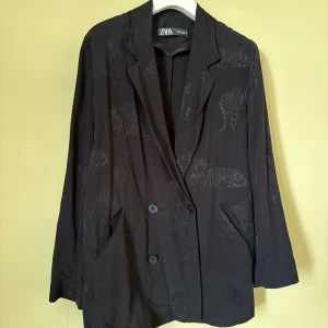 Svart kavaj med tigermönster från Zara - Svart kavaj från Zara med diskret broderade tigrar över hela plagget. Dubbelknäppt framtill, klassisk krage och två fickor. Materialet känns mjukt och har en snygg struktur. 