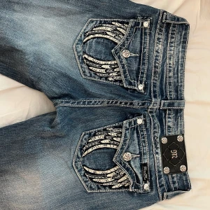 Miss Me blå low wais bootcut jeans med broderi - Säljer ett par blå Miss Me jeans i bootcut-modell med låg midja. Jeansen har snygga broderade detaljer och glittriga knappar på bakfickorna, samt slitningar och kontrastsömmar. Materialet är klassisk jeans med stretch för skön passform. Perfekt för dig som gillar statement-plagg!