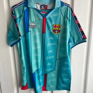 Barça Kappa Ronaldo 9 fotbollströja - Snygg Barcelona fotbollströja från Kappa med Ronaldo och nummer 9 på ryggen. Tröjan är turkosblå med röda och mörkblå detaljer, samt vita tryck. Har korta ärmar, krage och grafiskt mönster med Kappa-loggor och Barça-tryck. Perfekt för dig som älskar retrostil och fotboll.