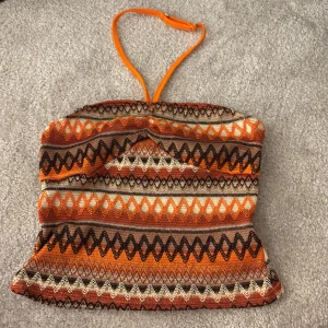 Mönstrad orange bandeau tubtopp - Säljer en färgstark bandeau tubtopp med justerbart orange axelband. Toppen har ett coolt zigzag-mönster i orange, brunt, beige och svart. Perfekt för dig som gillar retrovibbar och vill sticka ut med unika färger och mönster.