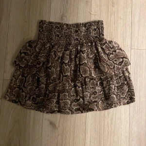 Brun kjol med ormmönster från Ü - Supersnygg kort kjol från Ü med coolt ormmönster i bruna och beige toner. Kjolen har bred smock i midjan och volanger som ger extra volym och attityd. Perfekt för dig som vill sticka ut med ett djuriskt print.