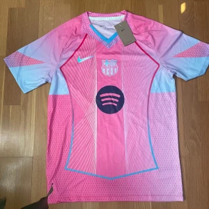 Barça x Nike 25/26 Special Edition Powder tröja - Snygg FC Barcelona fotbollströja från Nike i rosa och ljusblå toner med vita linjer och svart Spotify-logga på bröstet. V-ringad krage med blå kant, klubbmärke och Nike-logga framtill. 