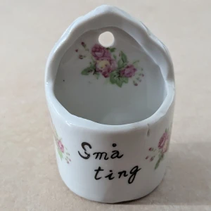 Liten porslinsbehållare - Liten porslinsbehållare med texten 'Små ting' och blommönster. Kan hängas på vägen eller ha stående på hylla eller bord eller liknade och används till att förvara små ting. En mindre defekt finns ett litet hack, defekten visas på bild