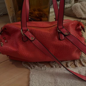 Röd axelväska med nitar från SMF - Cool röd axelväska i syntetmaterial med guldiga nitar på sidorna och flera dragkedjor. Väskan har en strukturerad yta och långa handtag med svarta kanter. Väskan har även massor med fickor inuti.