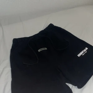 Svarta Essentials shorts Fear of God - Svarta shorts från Essentials Fear of God i storlek S. De har en loose passform, elastisk midja med snörning och en bakficka. Materialet är mjuk bomull, perfekt för chill dagar. Loggan är tryckt framtill i vitt för en clean vibe.