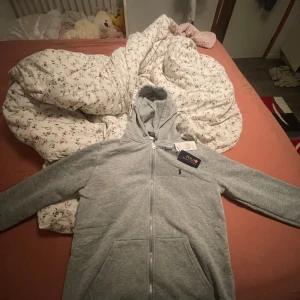 Grå hoodie från Polo Ralph Lauren - Säljer en grå hoodie från Polo Ralph Lauren med dragkedja och huva. Tröjan har långa ärmar, snörning vid huvan och klassisk Polo-logga broderad på bröstet. Perfekt för chill dagar och enkel att matcha med jeans eller joggers.