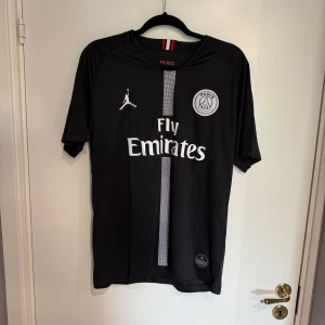 PSG x Jordan fotbollströja M - Snygg svart PSG fotbollströja i samarbete med Jordan. Tröjan har korta ärmar, vit logga och emblem, samt en vertikal randig detalj på framsidan. Materialet är lätt och andas, perfekt för match eller träning. Fly Emirates tryck på bröstet och Paris Saint-Germain badge.