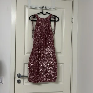 Rosa glittrig paljettklänning - Säljer en kort rosa klänning täckt med glittriga paljetter. Klänningen snygga kedjedetaljer över axlarna och en rund halsringning. Perfekt för dig som vill sticka ut och glänsa på nästa fest! Materialet är syntet med paljetter för extra bling. Har inte använt den en ända gång eftersom den var lite för liten för mig 