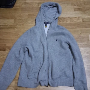 Grå hoodie från Polo Ralph Lauren - Säljer en chill grå hoodie från Polo Ralph Lauren med dragkedja och huva. Tröjan har lång ärm, klassisk Polo-logga på bröstet och är gjord i mjuk bomull. Perfekt för att slänga på sig när det är lite kyligt ute. Passar dig som gillar enkel och stilren streetwear.