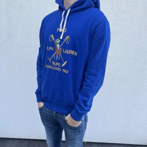 Blå hoodie från Polo Ralph Lauren - Säljer en blå hoodie från Polo Ralph Lauren med broderad polospelare och text på bröstet. Tröjan har huva med vita snören och ribbade muddar vid ärmar och nederkant. 