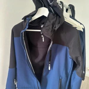 Blå och svart vindjacka med huva - Snygg vindjacka från Lindex i blått och svart med huva och vita detaljer. Jackan har dragkedjor på fickorna och bröstet, samt ett vitt kors på huvan och en vit V-detalj på ryggen. Perfekt för dig som gillar funktionell och stilren design.