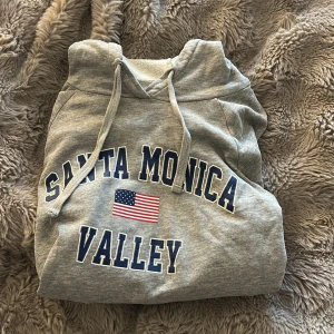 Grå hoodie Santa Monica Valley Gina Tricot - Grå hoodie från Gina Tricot med tryck 'Santa Monica Valley' och amerikansk flagga på bröstet. Tröjan har huva med snörning och är tillverkad i mjuk bomullsmix. Perfekt för chill dagar och streetstyle. Snyggt blått och vitt tryck framtill.