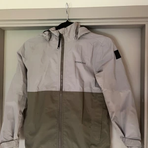 Grå och beige vindjacka Didriksons - Snygg tvåfärgad vindjacka från Didriksons i grått och beige. Jackan har huva, hel dragkedja framtill och stilren design med logga på bröstet. Perfekt för blåsiga dagar och enkel att matcha med jeans eller sneakers.