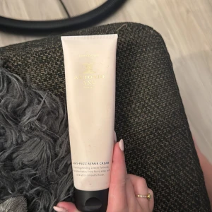 Antonio Axu Anti-Frizz Repair Cream - En ljusbeige tub med svart lock från Antonio Axu, innehållande Anti-Frizz Repair Cream. Perfekt för att ge håret en silkeslen och slät finish samtidigt som friss elimineras. Tuben har stilren design med guldtext och rymmer en stärkande krämformula.