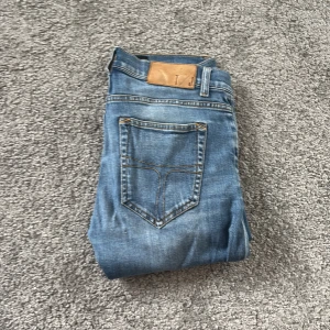 Blå jeans Tiger of Sweden 28/32 - Snygga blå jeans från Tiger of Sweden i klassisk denim. Modellen har fem fickor, ljusblå tvätt och kontrastsömmar. Passar både killar och tjejer och har en stilren look med knapp och dragkedja framtill. Perfekt för dig som gillar tidlös och enkel stil.