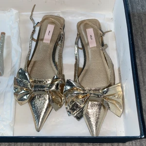 Guldiga ballerinaskor med rosett från Nelly - Säljer ett par glittriga ballerinaskor i guld från Nelly med spetsig tå och stor rosett framtill. Skorna har smala remmar runt hälen och platt sula. Perfekta för dig som vill sticka ut med metallic och cool design.