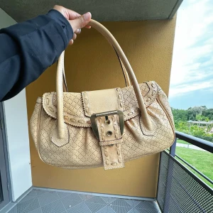 Vintage Beige handväska från Celine i skinn - Säljer en beige handväska från Celine i äkta skinn med snygga perforerade detaljer och ett stort spänne framtill. Väskan har två rejäla handtag och insida med dragkedja. Perfekt för dig som gillar klassisk design med coola detaljer.