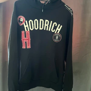 Svart hoodie från Hoodrich med tryck - Svart hoodie från Hoodrich med stora vita och röda tryck på framsidan, inklusive broderade märken och logotyp. Tröjan har huva med snörning och en klassisk känguruficka framtill. Perfekt för dig som gillar streetwear och vill sticka ut. Storlek M