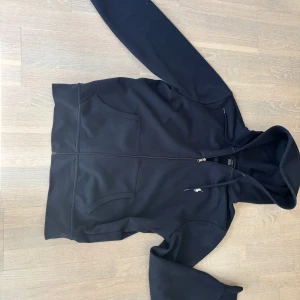 Svart hoodie från Polo Ralph Lauren - Svart hoodie från Polo Ralph Lauren med dragkedja och huva. Klassisk design med två fickor fram och den ikoniska broderade loggan på bröstet. Tillverkad i mjuk bomull, perfekt för chill dagar. Snygga detaljer som snörning i huvan och metalländar på snöret.