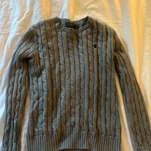 Grå kabelstickad tröja Polo Ralph Lauren - Säljer en aldrig använd grå kabelstickad tröja från Polo Ralph Lauren, med klassisk rund hals och broderad logga på bröstet. Tröjan har långa ärmar och ribbade muddar vid ärmslut och nederkant. Perfekt för dig som gillar stilren och tidlös design.