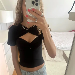 Svart croppad topp med cut-out - Säljer en svart croppad topp med snygg cut-out detalj över bröstet och korta ärmar. Toppen har en tight passform och är tillverkad i mjukt stretchigt material som sitter skönt mot kroppen. Perfekt för dig som gillar trendiga och unika plagg!