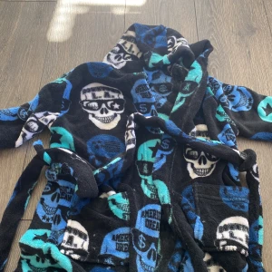 Morgonrock med döskallemönster Knox UWR - Säljer en mjuk och cool morgonrock Knox UWR i storlek 110/116. Västen har huva, fickor och bälte i midjan. Färgerna är svart, blå, turkos och vit med döskallemönster och texten 'American Dream'. Perfekt för dig som gillar edgy stil.