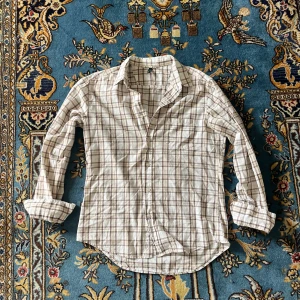 Rutig skjorta från Benetton - Snygg rutig skjorta från Benetton i beige och bruna toner. Klassisk krage, långärmad och med knappar framtill. Skjortan har en bröstficka och är gjord i mjuk bomull, perfekt för en chill och avslappnad stil.