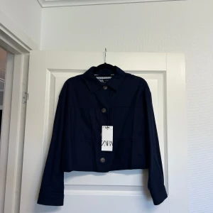 Mörkblå croppad overshirt från Zara - Snygg mörkblå croppad overshirt från Zara med klassisk krage och stora träknappar framtill. Jackan har långa ärmar med knappdetalj vid mudden och två stora fickor på bröstet. Materialet är linne, vilket ger en chill och lätt känsla.Ny med prislapp på. Aldrig använt bara provat. 