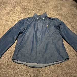 Blå skjorta från Ralph Lauren - Snygg blå skjorta från Ralph Lauren med klassisk krage och knappar framtill. Skjortan har lång ärm och ikonisk gul broderad logga på bröstet. Tillverkad i mjuk bomull som känns skön mot huden. Perfekt för dig som gillar stilrena och tidlösa plagg.