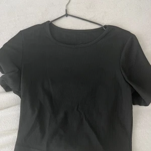 Svart ribbad topp med vit knyt - Säljer en svart ribbad topp med korta ärmar och rund hals. Baksidan har en öppen rygg med en stor vit knyt i chiffongliknande material som ger en snygg kontrast. Toppen är croppad och har en modern vibe, perfekt för dig som gillar coola detaljer.